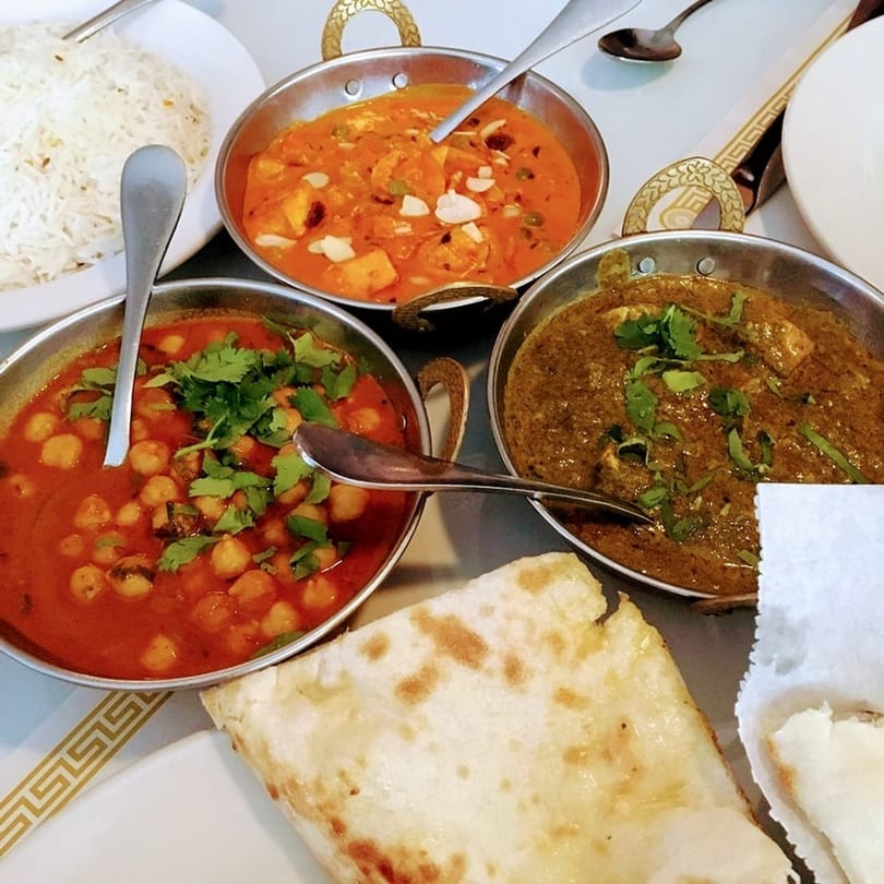 Three Indian curries in metal bowls with rice and naan bread on a white table.